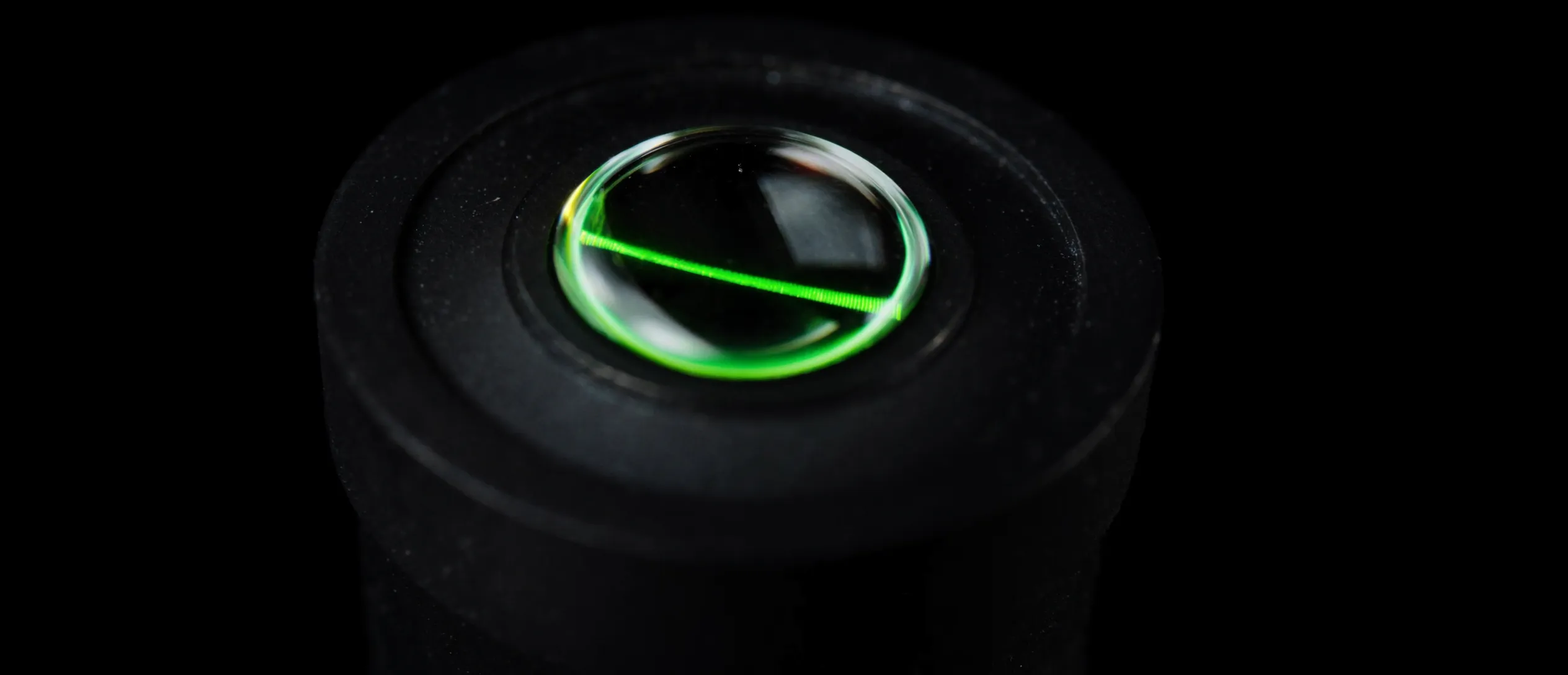 Macro of a precision lens element in matte black metal, a thin green laser refracting through its edge
